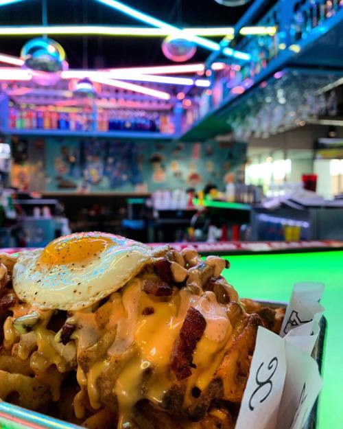 A vibrant bar scene with colorful neon lights, featuring a plate of loaded nachos topped with an egg in the foreground.