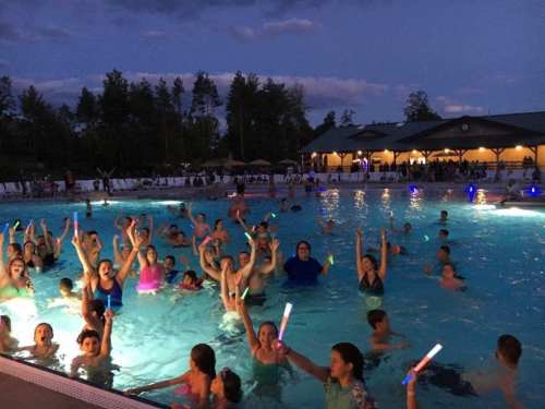 A lively group of children and teens enjoying a night swim at a pool, illuminated by colorful glow sticks.