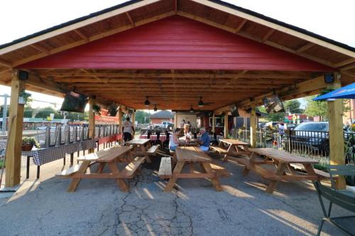 Outdoor dining area with wooden picnic tables under a covered pavilion, featuring fans and TVs, bustling with patrons.