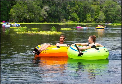 Two people relax on colorful inner tubes in a serene river, surrounded by lush greenery and other tubers in the background.