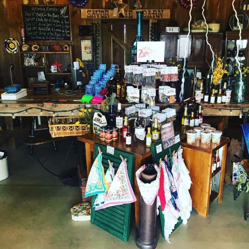 A rustic shop display featuring various bottles, jars, and colorful aprons in a warm, inviting barn setting.