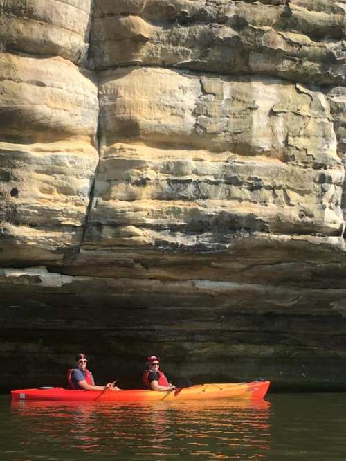 Two people in kayaks, one orange and one yellow, paddling under a rocky cliff on a calm waterway.