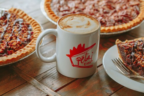 A mug of coffee sits on a wooden table next to two pecan pies, one on a plate with a fork.