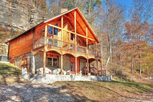 A rustic wooden cabin with a stone foundation, surrounded by trees and autumn foliage, on a sunny day.