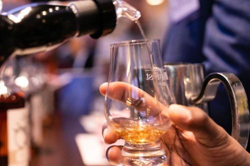 A hand pours whiskey into a glass at a tasting event, with blurred bottles in the background.