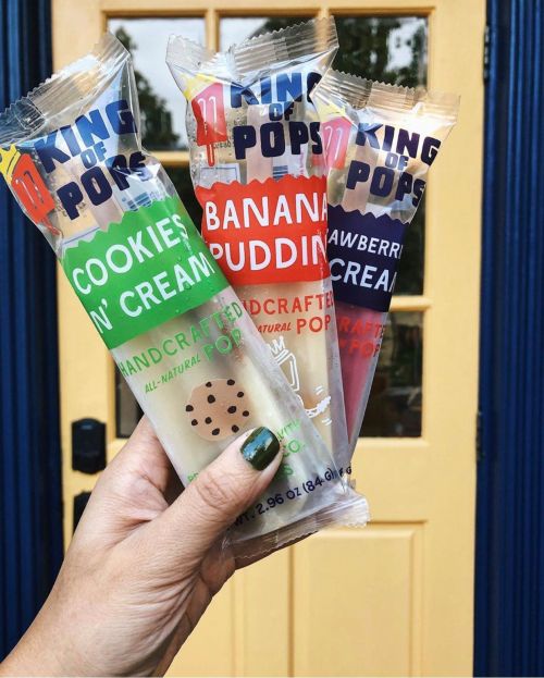 A hand holds three colorful ice pop packages: Cookies 'n' Cream, Banana Pudding, and Strawberry Cream, in front of a door.