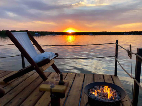 A serene lakeside scene at sunset, featuring a wooden deck, a chair, and a fire pit with glowing embers.