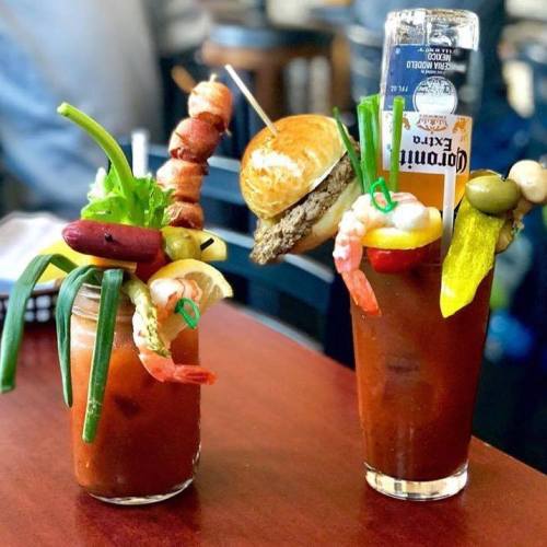 Two elaborate Bloody Mary cocktails garnished with shrimp, olives, pickles, and a mini burger, served with a beer.