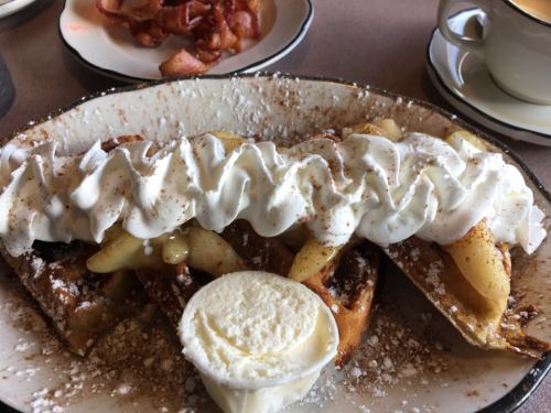 A plate of dessert crepes filled with apples, topped with whipped cream and a scoop of ice cream, with bacon in the background.