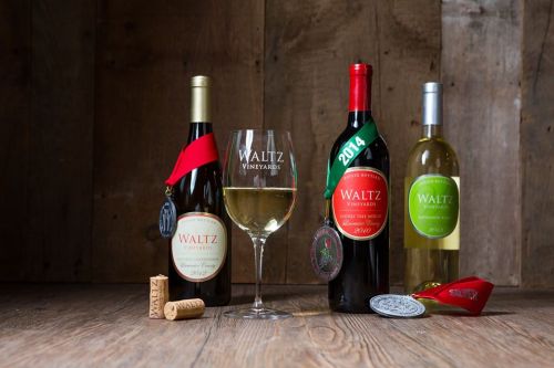 A glass of white wine with two bottles of Waltz Vineyards wine, corks, and medals on a wooden surface.