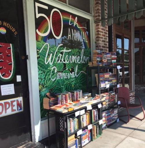 A colorful storefront with a mural for a watermelon carnival and a display of books on a table outside.