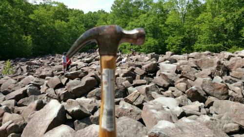 A close-up of a hammer in the foreground, with people exploring a rocky landscape in the background.