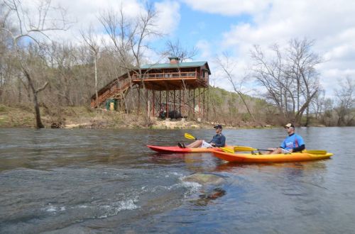 Two people kayaking on a river with a wooden cabin on stilts in the background, surrounded by trees and blue sky.
