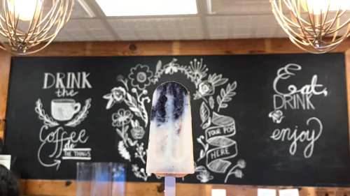 A popsicle in front of a chalkboard with floral designs and text promoting coffee and enjoyment.