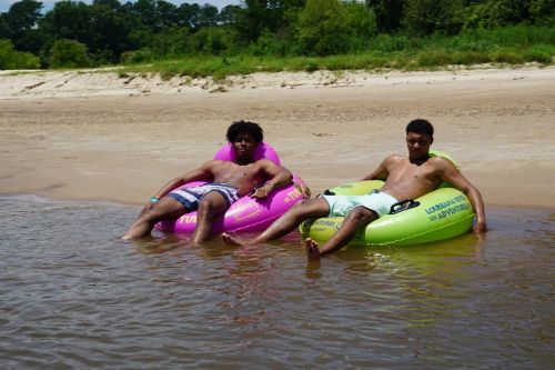 Two young men relax on colorful inner tubes in shallow water on a sandy beach, surrounded by greenery.
