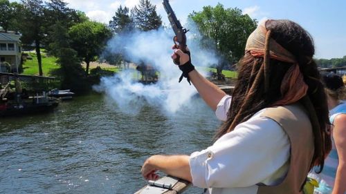 A person in pirate attire fires a pistol over a river, with smoke rising and trees in the background.