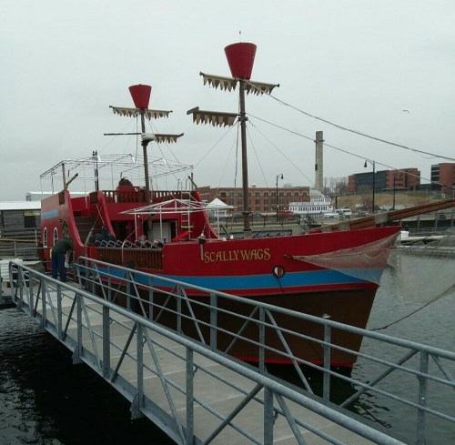 A colorful pirate-themed boat named "Scallywags" docked at a marina on a cloudy day.