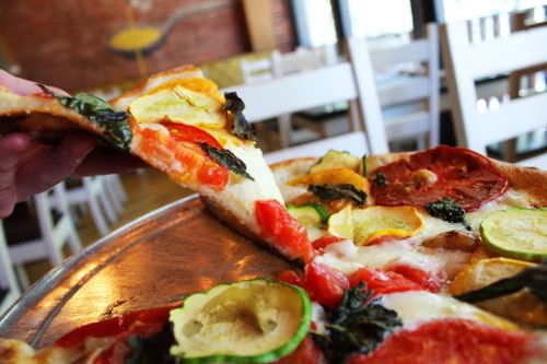A hand lifts a slice of pizza topped with tomatoes, zucchini, and basil from a metal tray in a cozy restaurant setting.