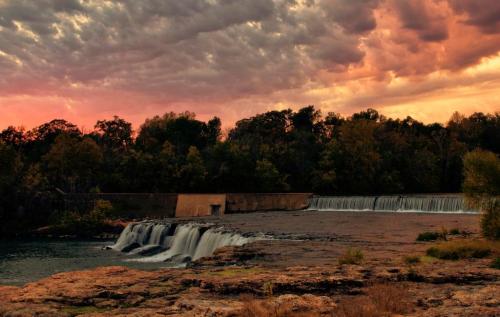 A serene waterfall cascades over rocks under a colorful sunset sky, surrounded by lush trees and clouds.