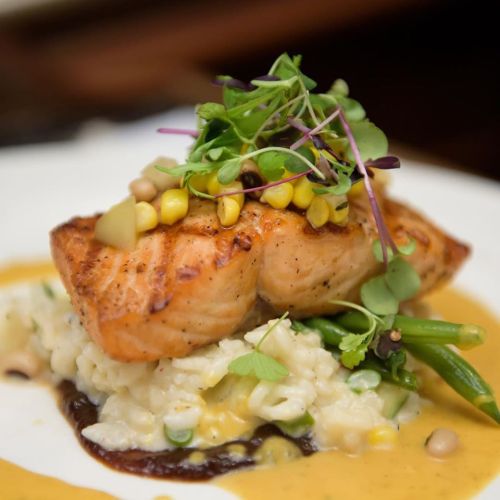 Grilled salmon atop creamy risotto, garnished with corn, microgreens, and a drizzle of sauce.