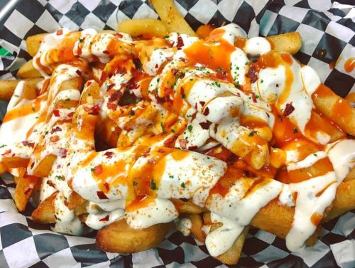 A basket of fries topped with spicy sauce, ranch dressing, and sprinkled with herbs and bacon bits.