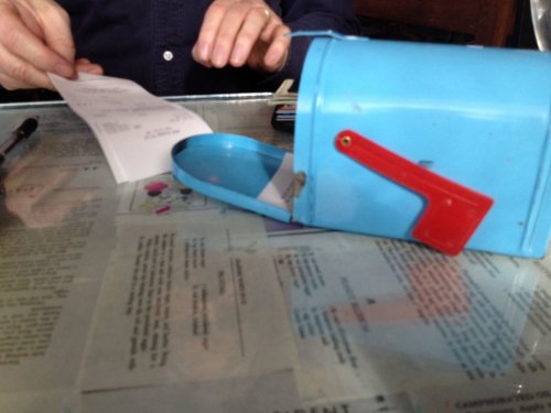 A blue mailbox with a red flag sits on a table, alongside a receipt and a hand holding a pen.