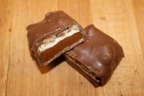 Two chocolate-covered candy bars, one cut in half to reveal a creamy filling and nuts inside, on a wooden surface.