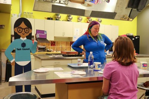A woman with colorful hair talks to a girl in a kitchen, with a cartoon figure of a girl in a blue shirt nearby.
