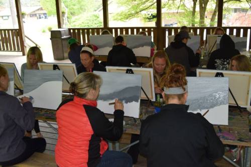 A group of people painting landscapes on canvases in a covered outdoor space.