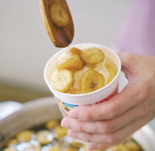 A hand holds a cup of ice cream topped with sliced bananas and a drizzle of syrup.