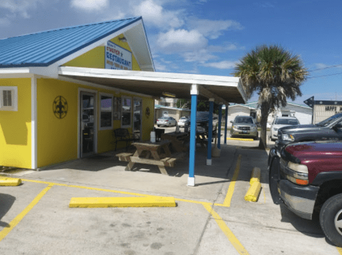 A bright yellow restaurant with a blue roof, outdoor seating, and parked cars in a sunny parking lot.