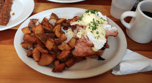 A plate of eggs Benedict with ham and hollandaise sauce, served with crispy potato wedges and a cup of coffee.