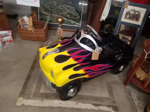 A small, vintage toy car painted yellow with black flames, displayed in an antique shop among various items.