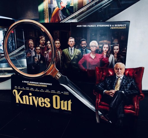 A promotional display for "Knives Out" featuring a large magnifying glass and characters from the film.