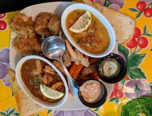 A colorful plate featuring fried shrimp, seafood gumbo, sweet potato fries, and dipping sauces, with lemon slices.