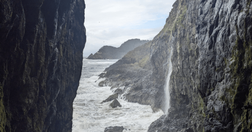A rocky coastal scene with a waterfall cascading into the ocean, framed by steep cliffs and a cloudy sky.