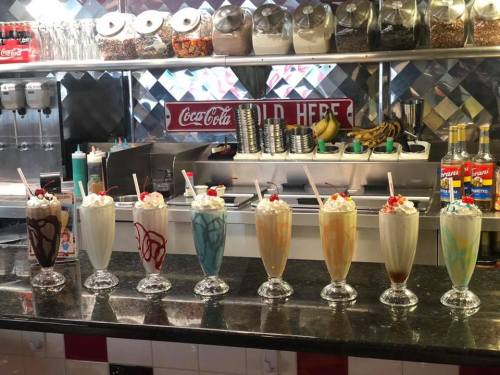 A row of colorful milkshakes topped with whipped cream and cherries, displayed in tall glasses at a retro diner.