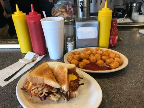A plate with a sandwich and tater tots, accompanied by condiments and a drink on a diner table.