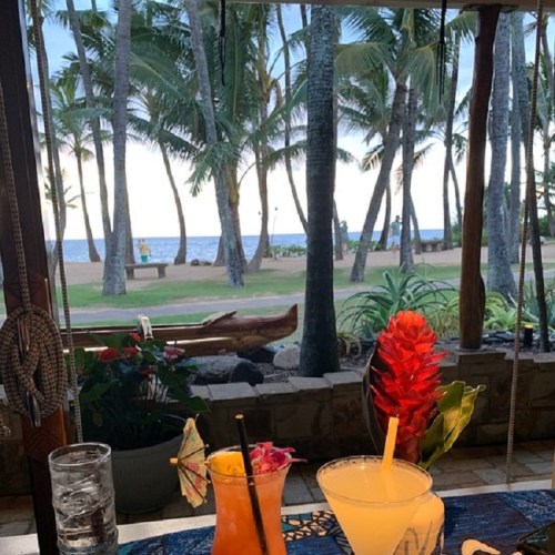 Tropical scene with two cocktails on a table, palm trees, and ocean view in the background.