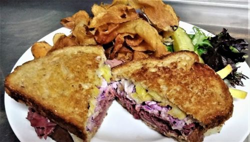 A sliced sandwich filled with meat and coleslaw, served with crispy potato chips and a side of mixed greens.
