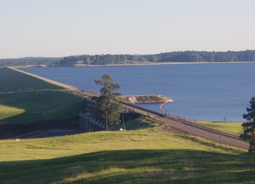 A scenic view of a dam with a road crossing over, surrounded by green hills and a calm blue lake.
