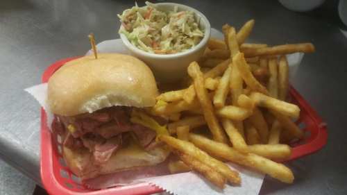 A plate featuring a sandwich with meat and mustard, a side of fries, and a small bowl of coleslaw.