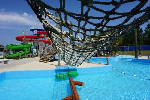 A water park scene featuring colorful slides, a rope net, and a blue pool with floating rings under a clear sky.