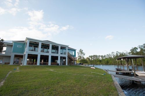 A large waterfront house with a deck, surrounded by green grass and trees, next to a calm river.