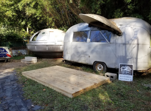 Two vintage trailers parked on grass, one rounded and one streamlined, with a wooden platform and a historic district sign nearby.