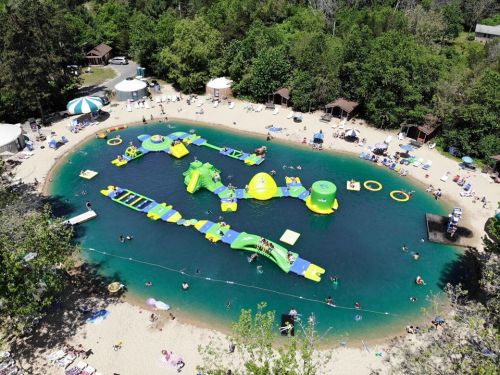 Aerial view of a sandy beach with inflatable water attractions and people enjoying the water in a green, wooded area.