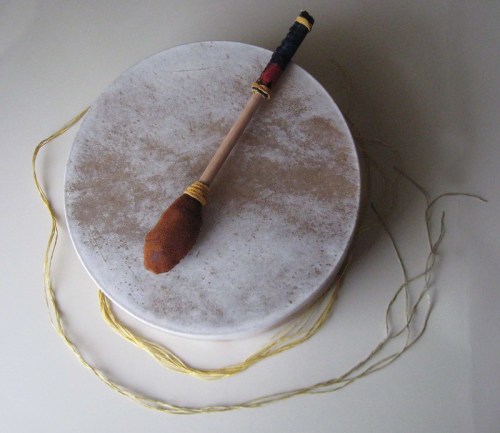 A round drum with a wooden mallet resting on top, surrounded by yellow string on a light background.