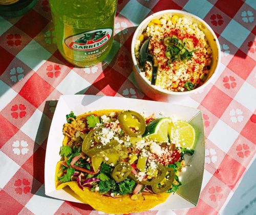 A vibrant taco topped with jalapeños and cheese, alongside a bowl of corn and a bottle of Jarritos on a checkered tablecloth.