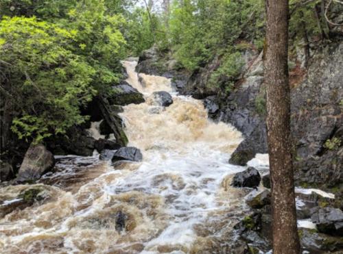 A rushing waterfall cascades over rocks, surrounded by lush green trees and a rocky landscape.