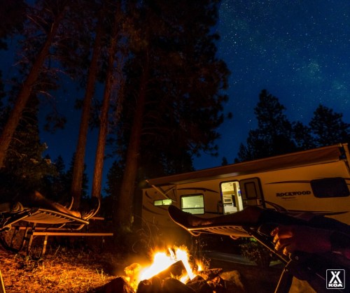 A cozy campsite at night with a campfire, two chairs, and a camper under a starry sky surrounded by trees.